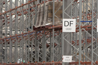 Warehouse workers in yellow safety vests push carts with boxes, surrounded by tall metal racks. A "DF ODD" sign hangs above them.