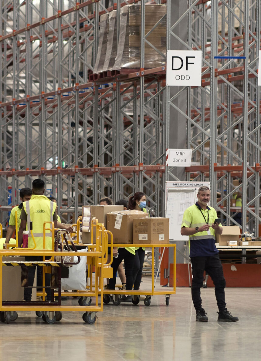 Warehouse workers in yellow safety vests push carts with boxes, surrounded by tall metal racks. A "DF ODD" sign hangs above them.