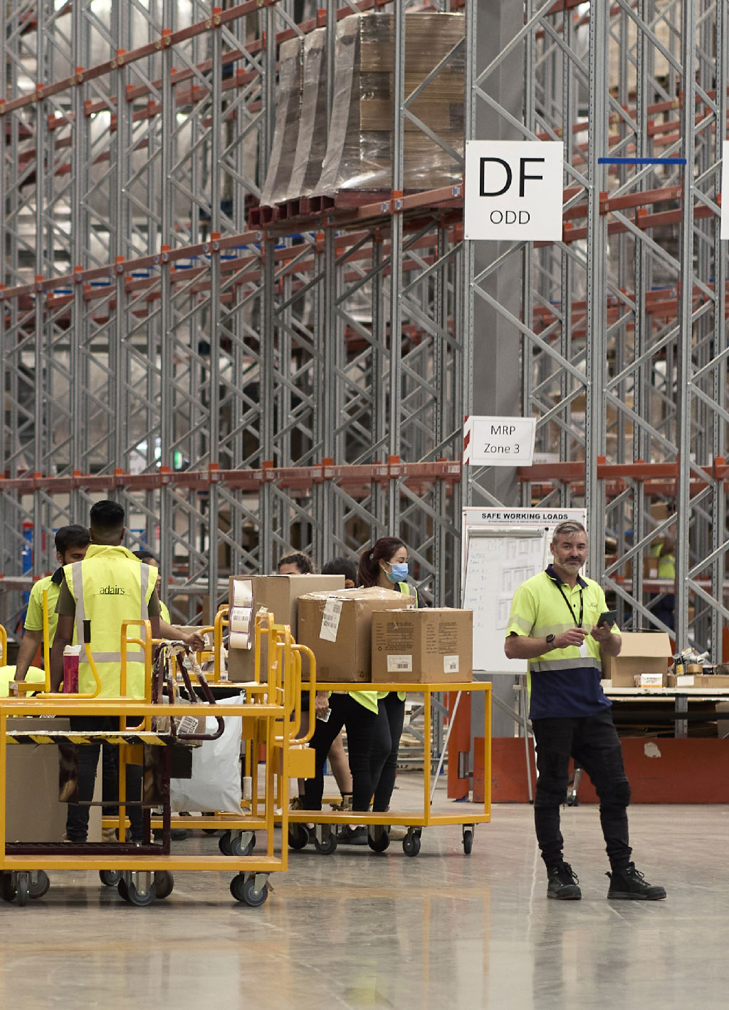 Warehouse workers in yellow safety vests push carts with boxes, surrounded by tall metal racks. A "DF ODD" sign hangs above them.