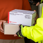 A person in a bright yellow jacket hands over two Australia Post parcels to another person wearing a red sweater. The scene conveys efficient delivery.