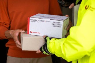 A person in a bright yellow jacket hands over two Australia Post parcels to another person wearing a red sweater. The scene conveys efficient delivery.