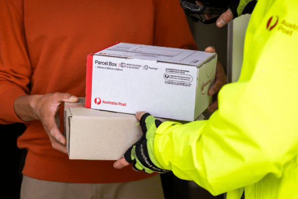 A person in a bright yellow jacket hands over two Australia Post parcels to another person wearing a red sweater. The scene conveys efficient delivery.