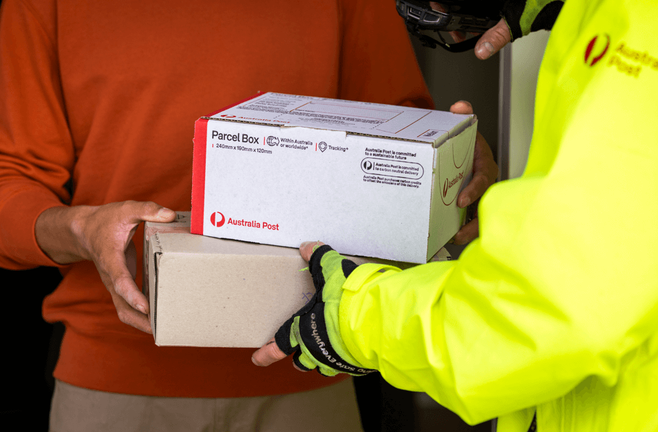 A person in a bright yellow jacket hands over two Australia Post parcels to another person wearing a red sweater. The scene conveys efficient delivery.