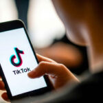 A person holds a smartphone displaying the TikTok logo on the screen. The setting is indoor with a soft background, creating a focused and casual mood.