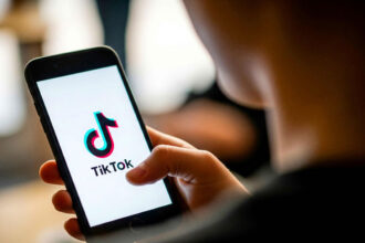 A person holds a smartphone displaying the TikTok logo on the screen. The setting is indoor with a soft background, creating a focused and casual mood.