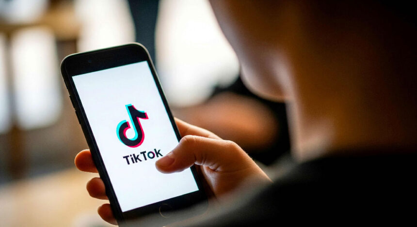 A person holds a smartphone displaying the TikTok logo on the screen. The setting is indoor with a soft background, creating a focused and casual mood.