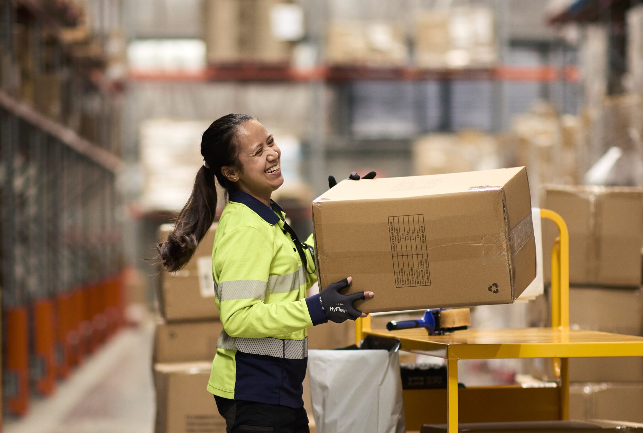 Warehouse worker in a high-visibility uniform smiling while lifting a large cardboard box beside a packing station, with shelving and stacked cartons in the background of a busy distribution centre.