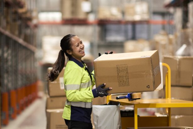 Warehouse worker in a high-visibility uniform smiling while lifting a large cardboard box beside a packing station, with shelving and stacked cartons in the background of a busy distribution centre.