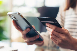 A person in a striped shirt holds a smartphone and a credit card, appearing to make an online purchase. The scene conveys modern technology and convenience.