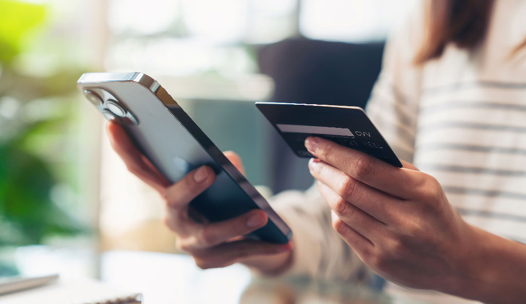 A person in a striped shirt holds a smartphone and a credit card, appearing to make an online purchase. The scene conveys modern technology and convenience.