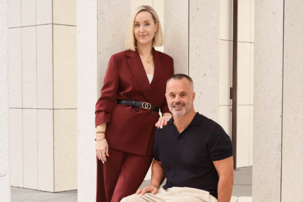Joanna Robinson, CMO, stands beside Joshua Nunan, Director of Platform Media & Services, as they pose together in a modern office setting with stone columns. Joanna rests a hand on Joshua's shoulder while both smile at the camera