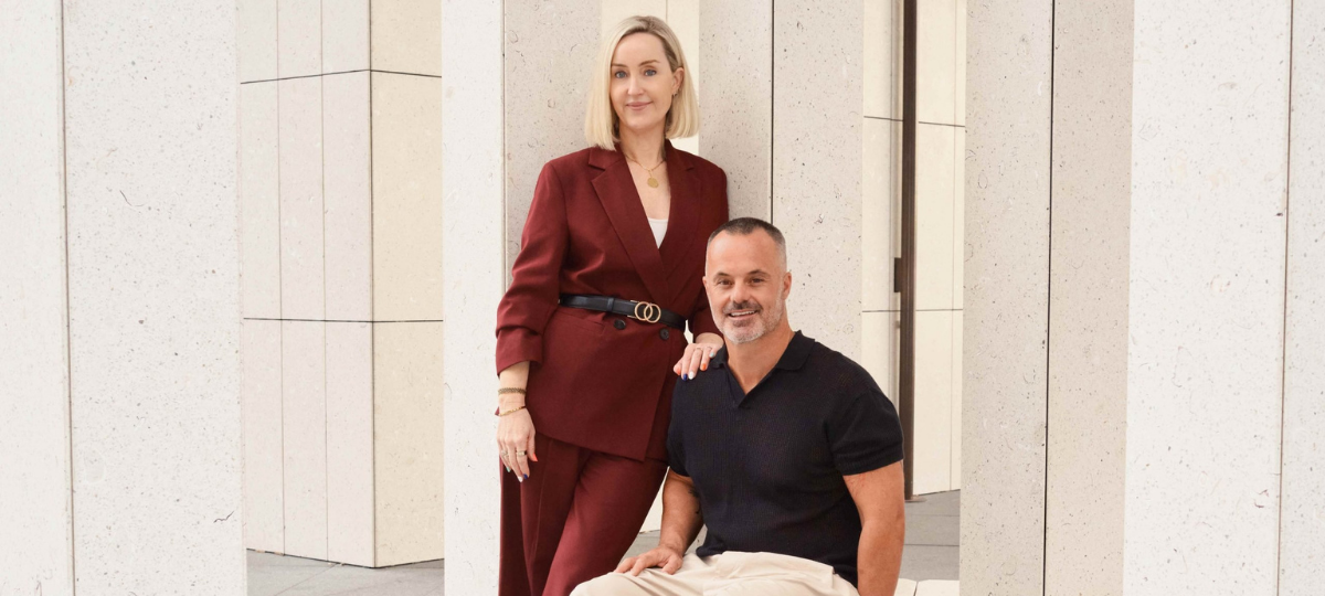 Joanna Robinson, CMO, stands beside Joshua Nunan, Director of Platform Media & Services, as they pose together in a modern office setting with stone columns. Joanna rests a hand on Joshua's shoulder while both smile at the camera
