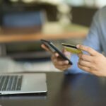 A person holds a credit card and smartphone while sitting at a desk with an open laptop, making an online purchase or digital payment.
