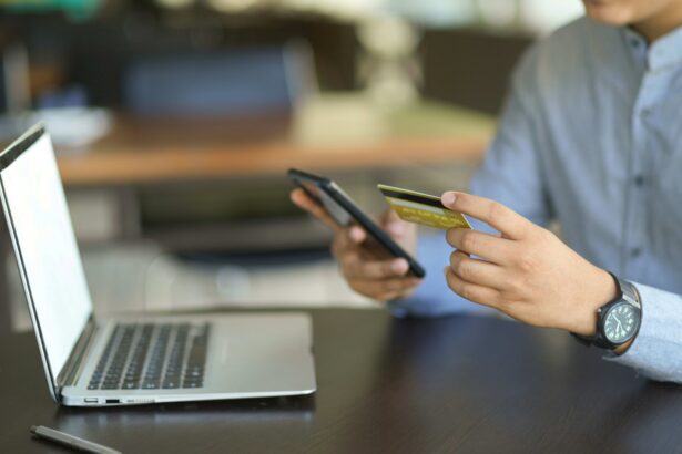 A person holds a credit card and smartphone while sitting at a desk with an open laptop, making an online purchase or digital payment.