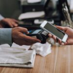 A customer holds a smartphone near a payment terminal to make a contactless payment at a retail checkout counter.