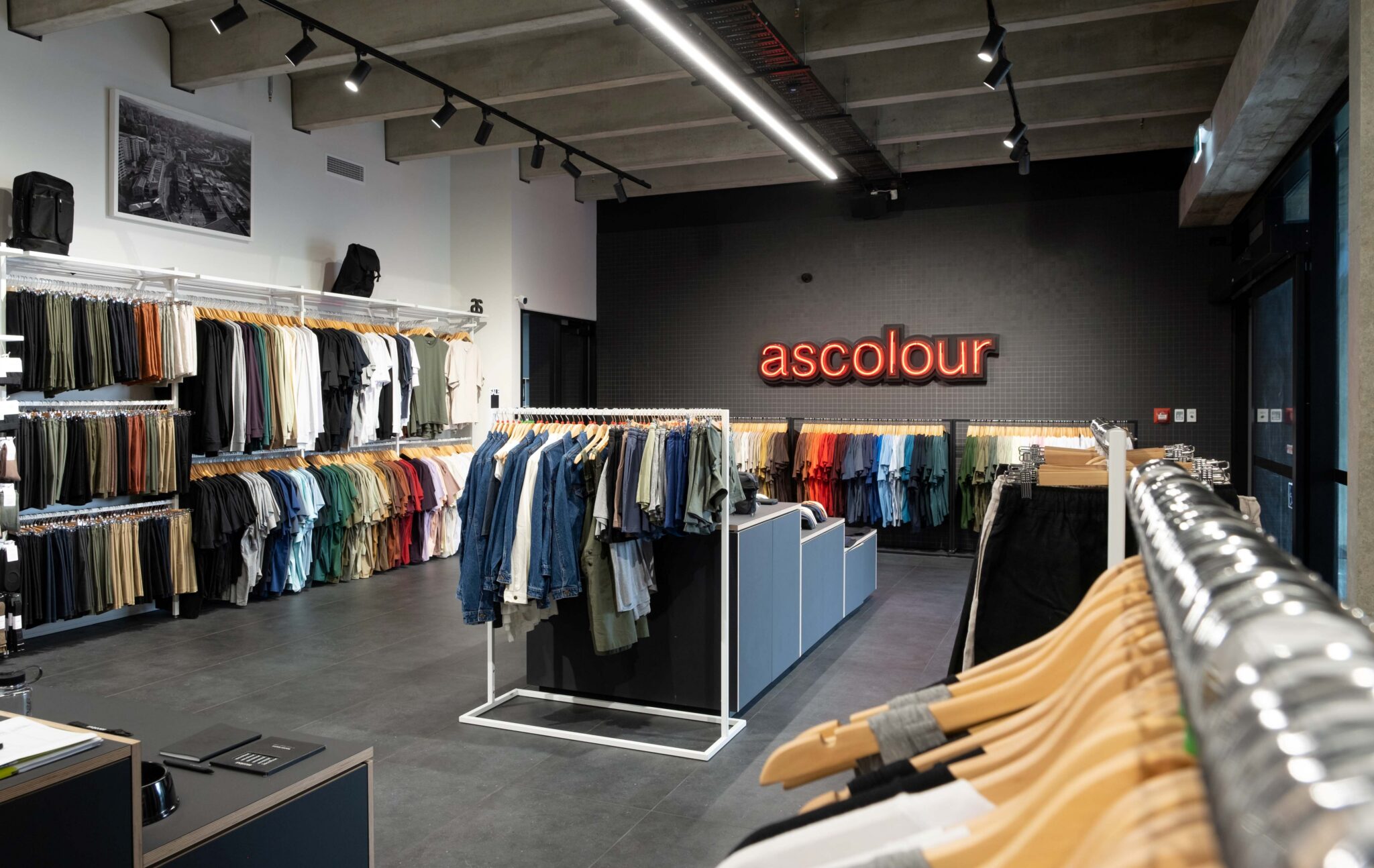 Interior of an AS Colour retail store with neatly organised racks of T-shirts, denim and basics arranged by colour. A red illuminated "ascolour" sign is displayed on the back wall, highlighting the brand's minimalist retail fit-out.