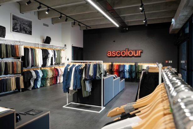 Interior of an AS Colour retail store with neatly organised racks of T-shirts, denim and basics arranged by colour. A red illuminated "ascolour" sign is displayed on the back wall, highlighting the brand's minimalist retail fit-out.