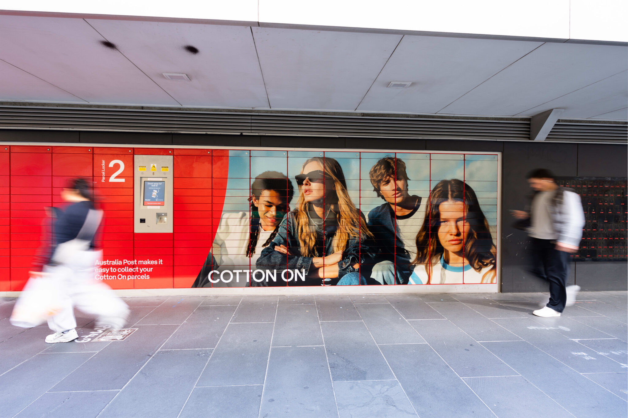 Australia Post parcel lockers featuring Cotton On advertising, with pedestrians walking past.