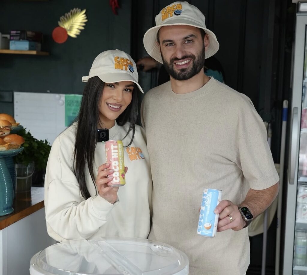 Two people wearing Coco Hit branded hats pose together while holding cans of Coco Hit coconut water at a promotional event.
