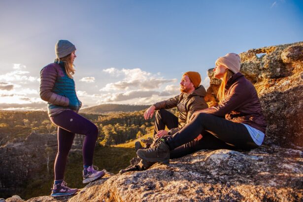 Three hikers wearing insulated outdoor clothing rest on a rocky mountain lookout at sunset, overlooking a rugged bush landscape, representing outdoor adventure, hiking and nature exploration.