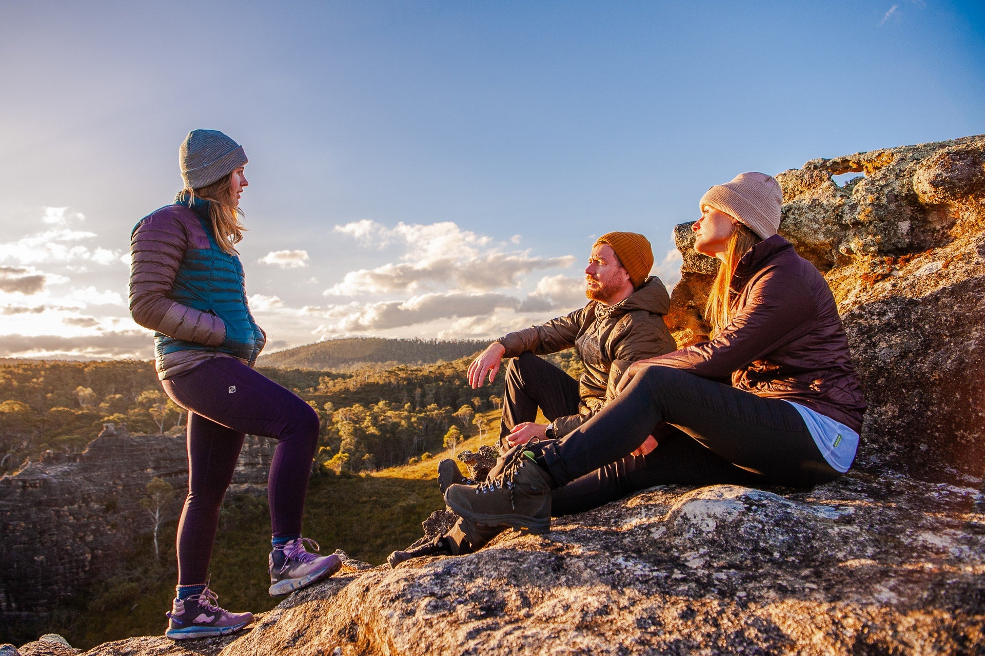 Three hikers wearing insulated outdoor clothing rest on a rocky mountain lookout at sunset, overlooking a rugged bush landscape, representing outdoor adventure, hiking and nature exploration.
