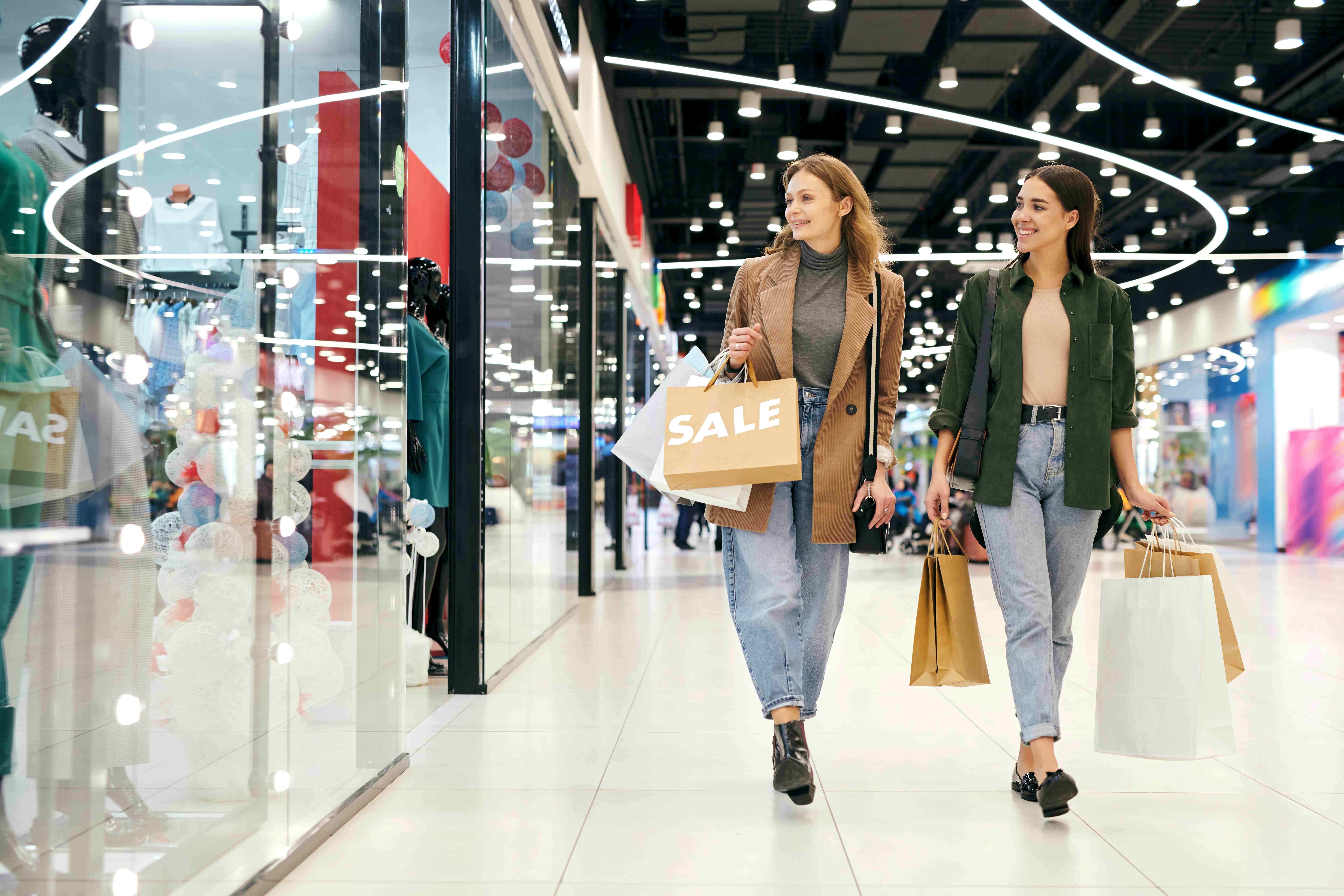 Two shoppers carrying shopping bags walk through a brightly lit shopping centre during a retail sale.
