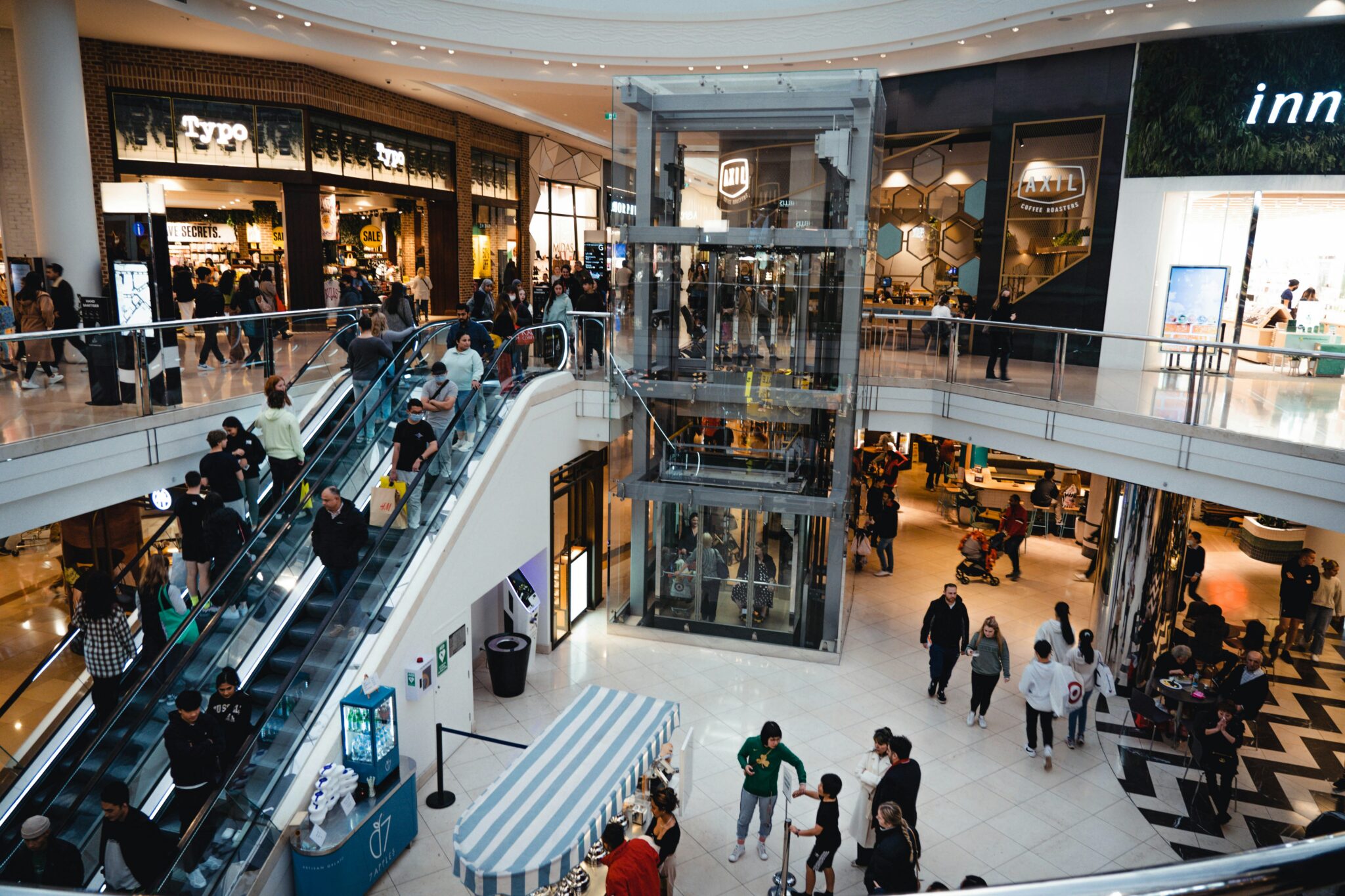 Shoppers move between stores, escalators and cafés across multiple levels of a busy shopping centre.