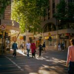 Pedestrians crossing a busy Melbourne CBD street in afternoon sunlight, surrounded by shops and city buildings.