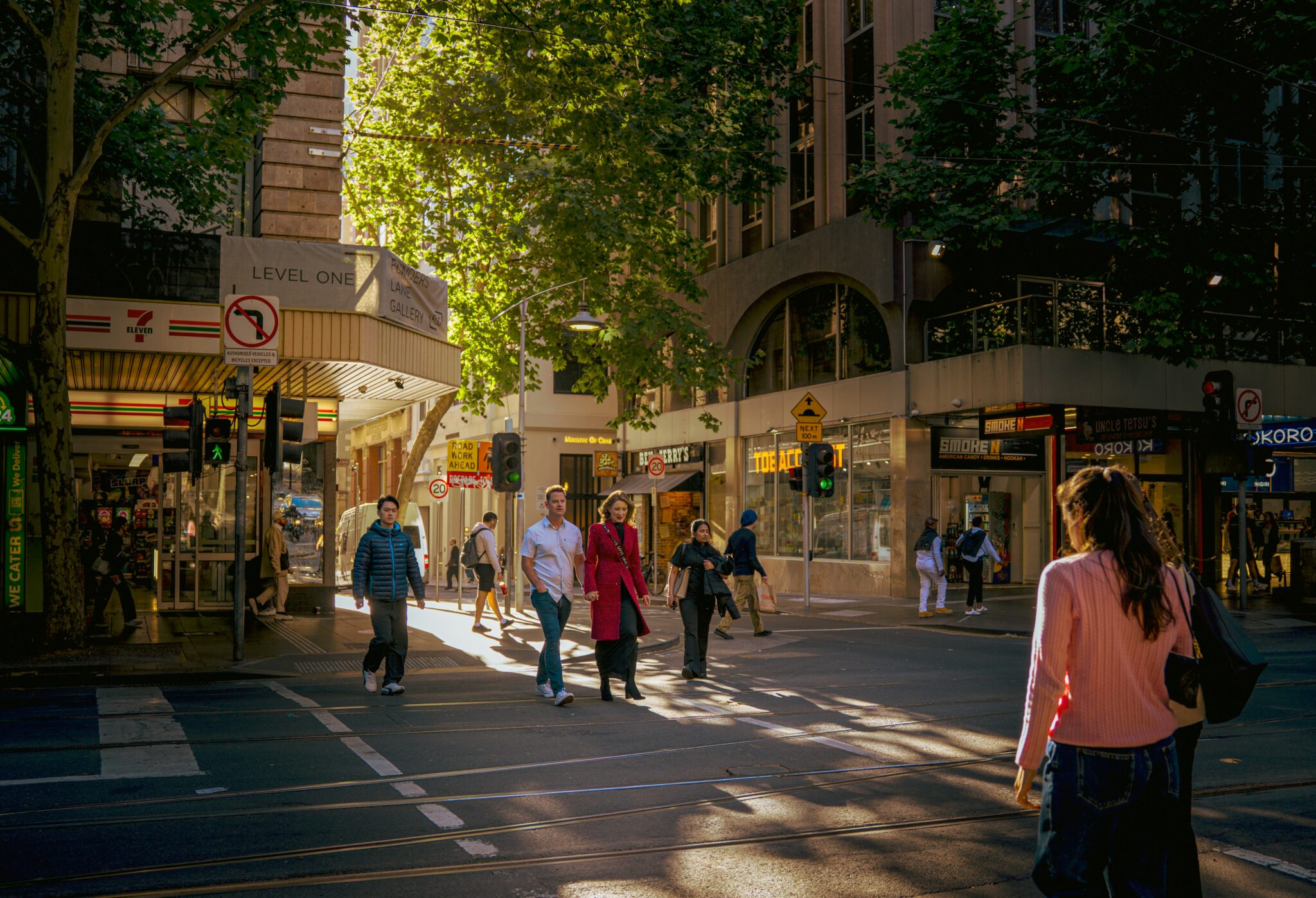 Pedestrians crossing a busy Melbourne CBD street in afternoon sunlight, surrounded by shops and city buildings.