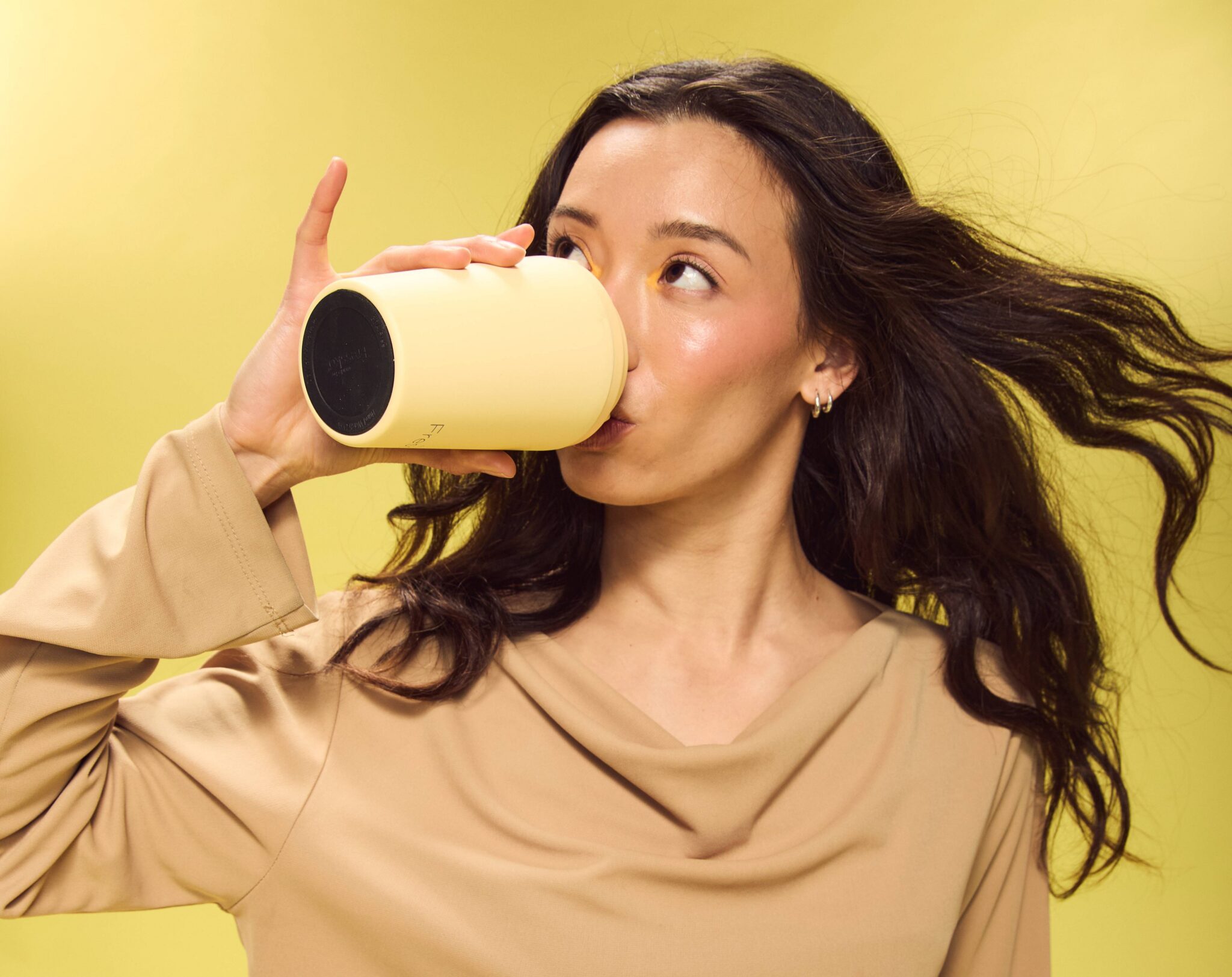 Woman drinking from a pale yellow made by Fressko reusable coffee cup against a bright yellow background, highlighting the Melbourne-based sustainable drinkware brand.