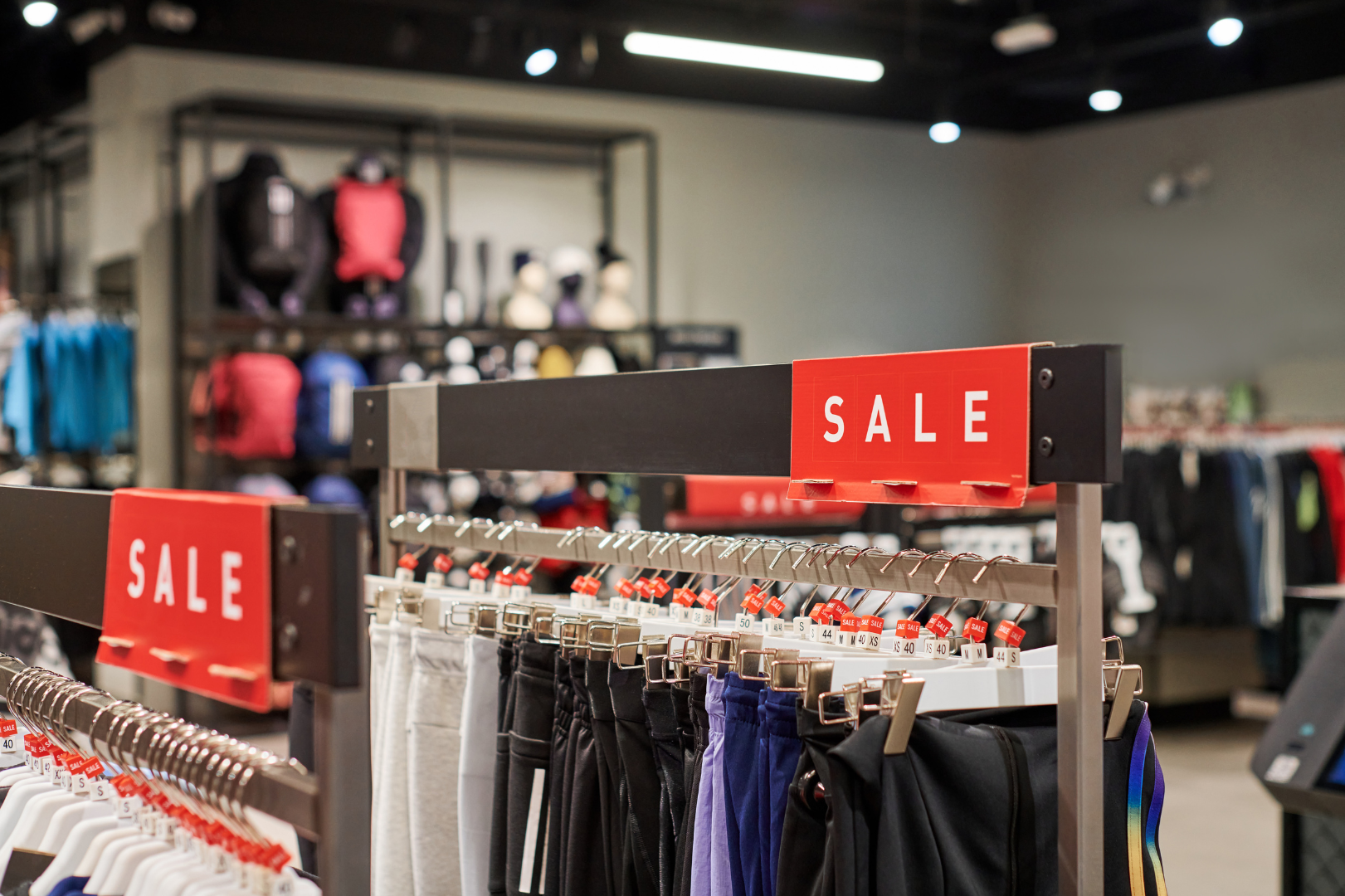 Clothing racks displaying discounted sportswear beneath red sale signs inside a retail store.